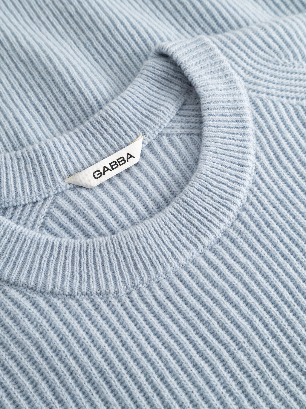 Close-up of a dusty blue ribbed crew neck sweater, the GABremy O-Neck Flash LS Knit by Gabba, featuring a white clothing tag sewn at the neckline with the Gabba brand name in black letters. The image highlights the collar and the texture of the 100% wool fabric.