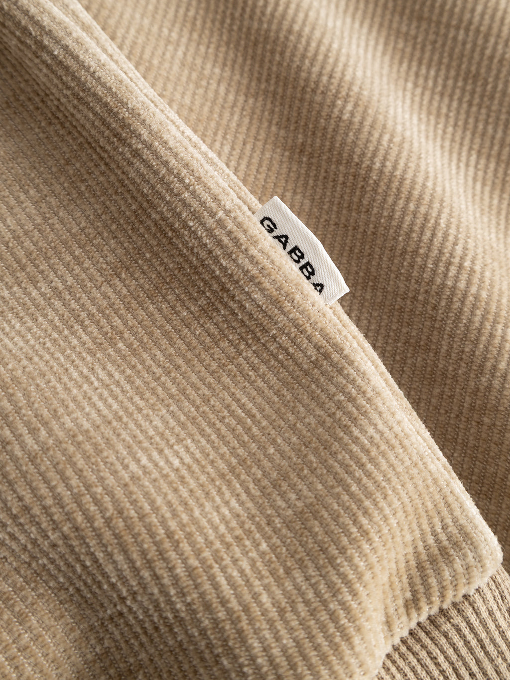 Close-up of the beige GABprimo Fune Hoodie LS Sweat - Sand Crocker by Gabba, showing its jersey corduroy fabric and a small white tag labeled GABBA sewn into the seam.