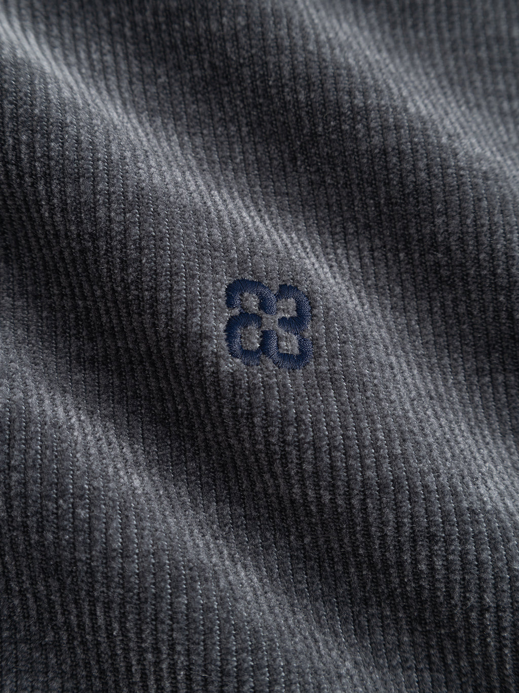 Close-up of the ribbed gray jersey corduroy fabric from the Gabba GABprimo Fune Hoodie LS Sweat in Turbulence Blue, featuring a small, dark blue embroidered 83 near the center. The lighting accentuates the texture and folds of the material, highlighting its cozy, casual appeal and regular fit silhouette.