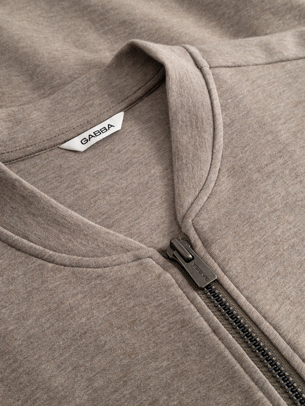 Close-up of the GABnik Camp Lock Sweat - Sand Mel. by Gabba, a beige zip-up sweatshirt featuring a bomber collar, black zipper, and a white clothing label with the Gabba brand name inside the collar. Crafted from mid-weight interlock fabric, this modern outerwear piece combines style and comfort.