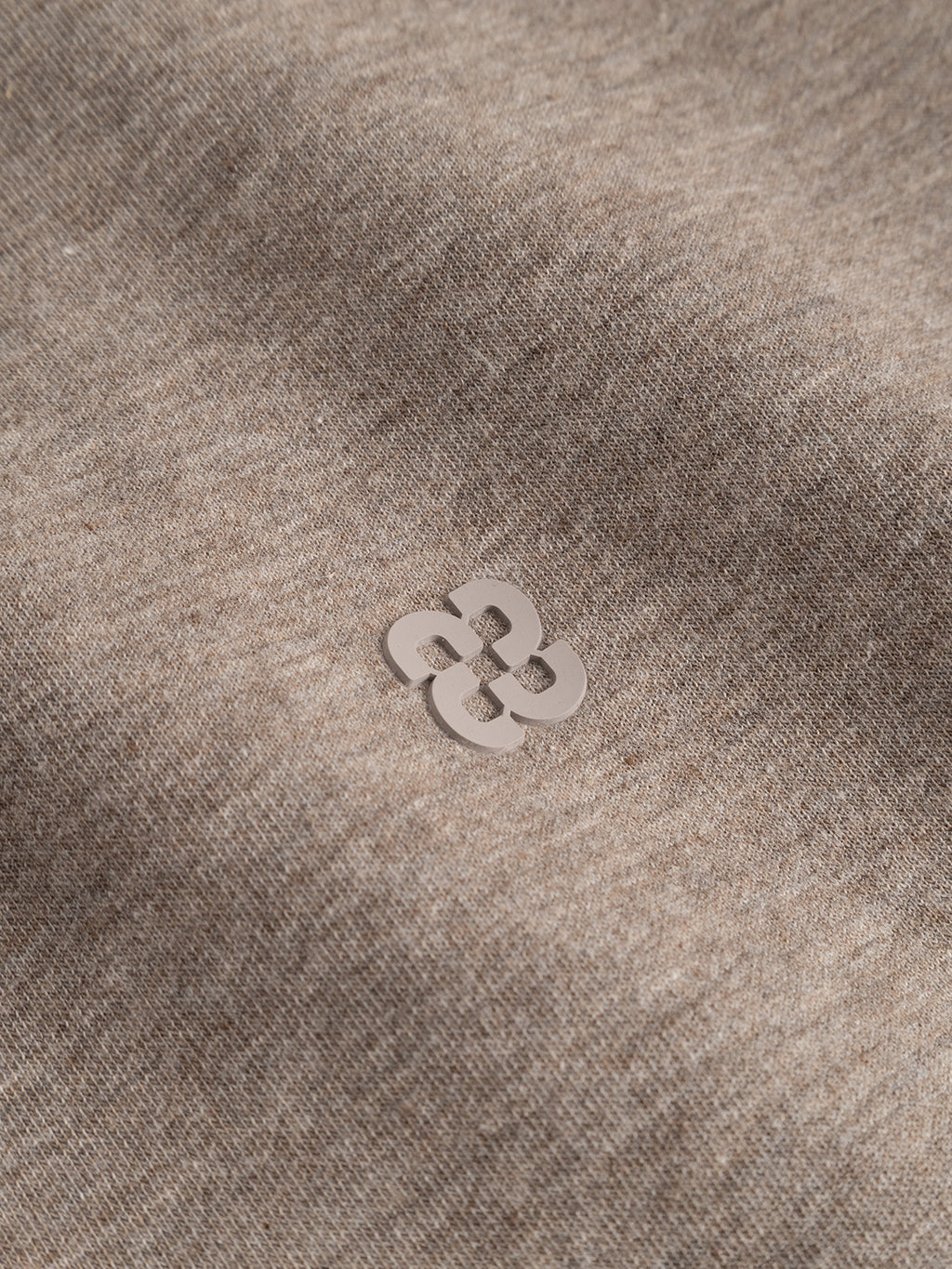 A small, light-colored abstract flower-shaped pin is placed on the GABnik Camp Lock Sweat – Sand Mel. by Gabba, a piece of modern outerwear in brown mid-weight interlock fabric, with soft natural lighting and gentle shadows highlighting its textured finish.