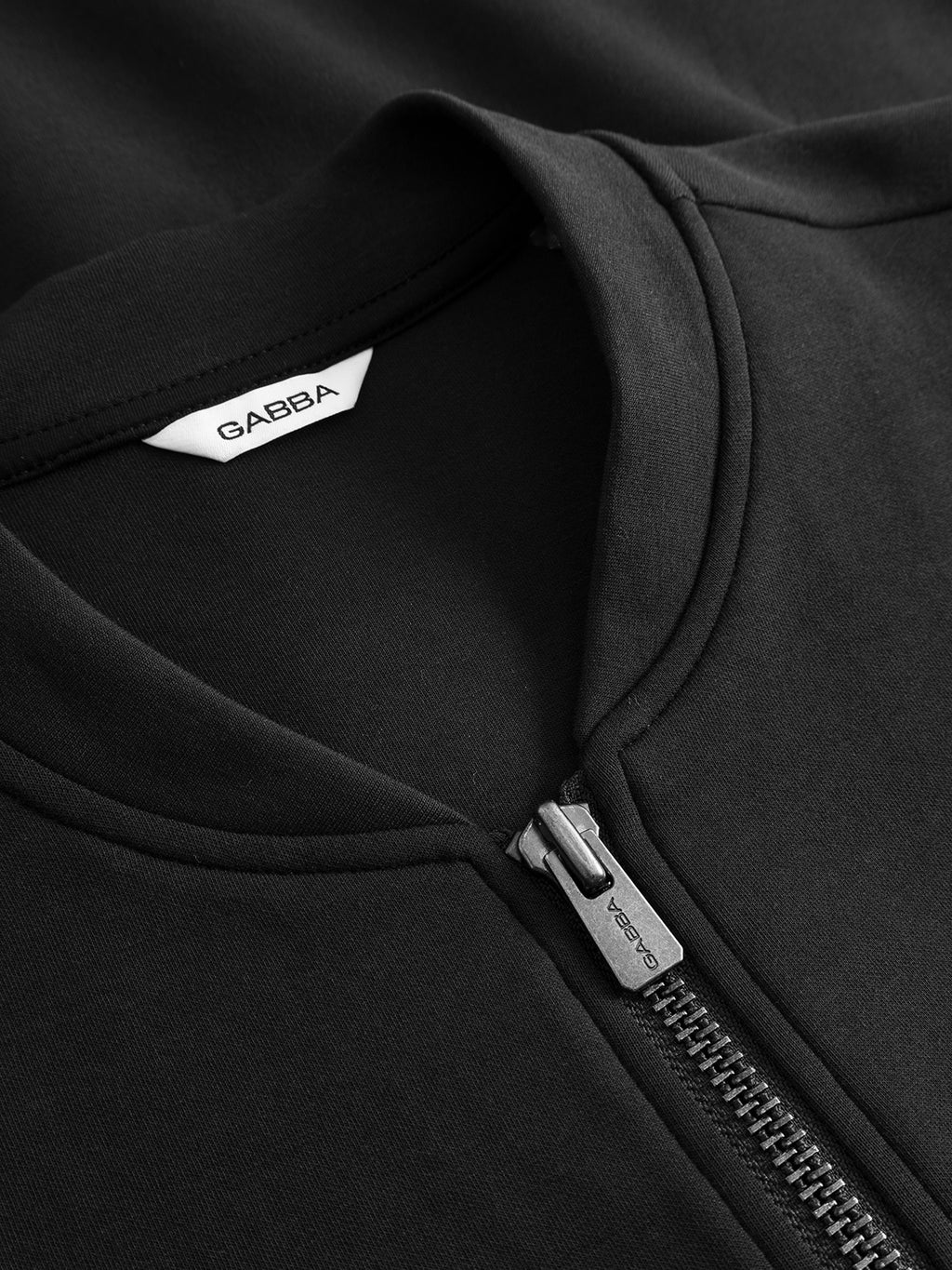 Close-up of the Gabba GABnik Camp Lock Sweat - Meteorite, a black zip-up garment featuring a visible silver zipper and a white GABBA label inside the collar. The mid-weight interlock fabric appears smooth and expertly stitched, embodying the precise craftsmanship typical of modern outerwear.