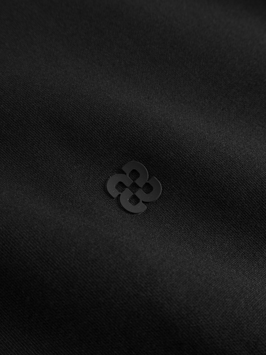 A close-up of the meteorite black mid-weight interlock fabric of the Gabba GABnik Camp Lock Sweat, featuring a small, dark geometric logo printed in the center. The logo consists of four rounded shapes forming a symmetrical pattern—a refined detail that elevates modern outerwear such as bomber collar jackets.