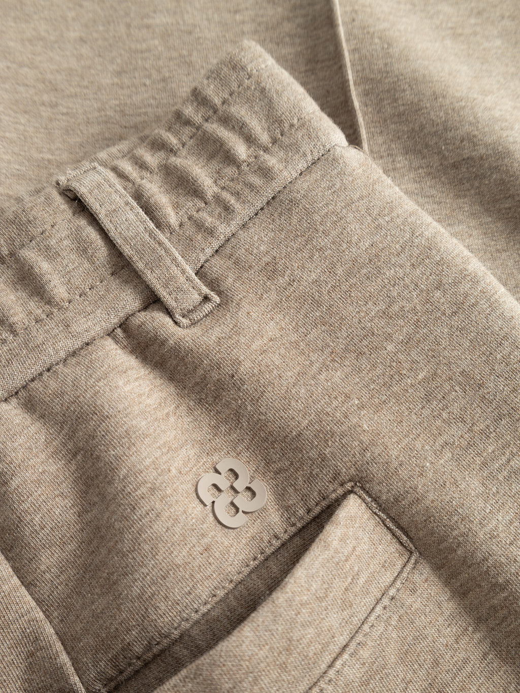 Close-up of the Gabba GABmonza Cropped Tasca Lock Pant in Sand Mel showcases its elastic waistband, belt loop, and flat pocket. Near the pocket opening is a small, white, clover-shaped logo. Crafted from mid-weight interlock fabric for a relaxed straight fit, the pants feature a soft and slightly textured appearance.