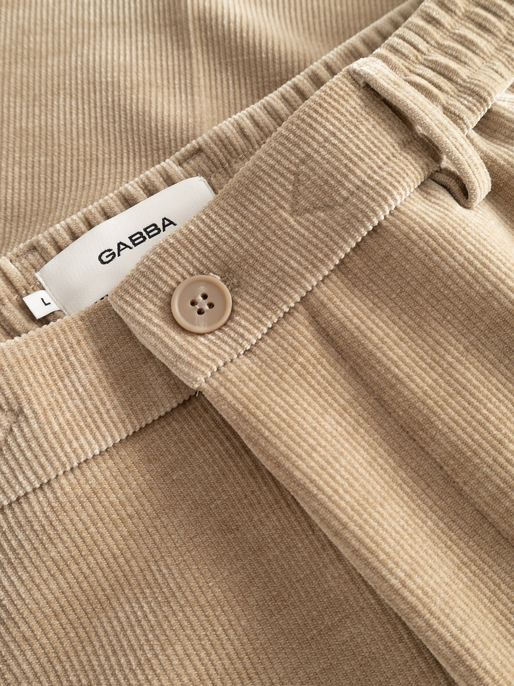 Close-up of the Gabba GABmonza Cropped Tasca Fune Pant in Sand Crocker, featuring textured beige corduroy fabric, a button closure, belt loops, and a visible white label reading “Gabba.” The relaxed straight fit is accentuated by the detailed stitching and tactile material.