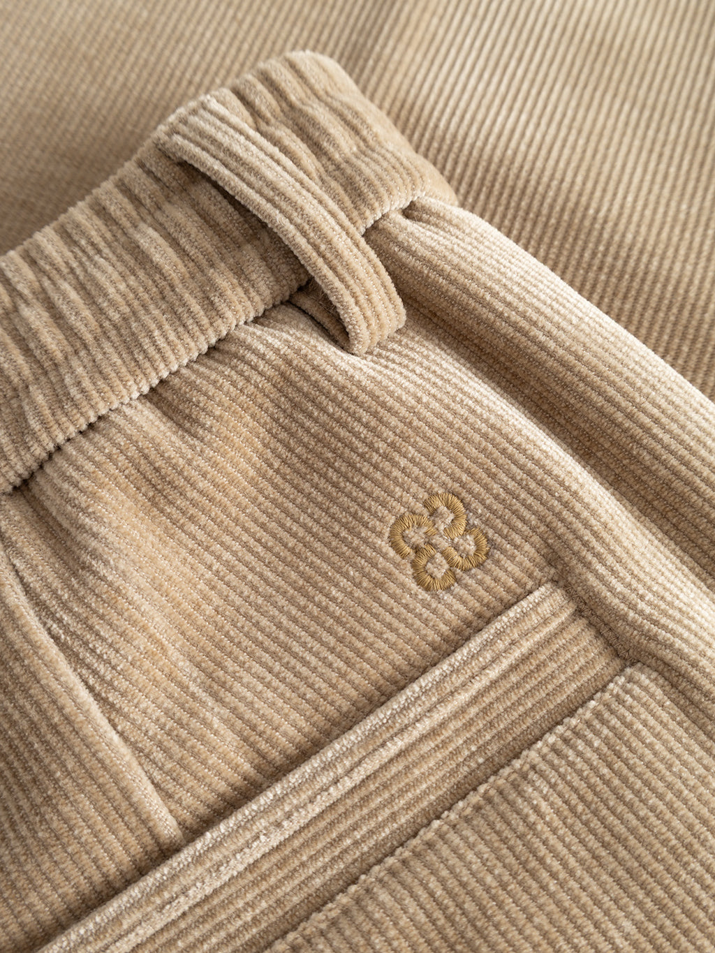 Close-up of the Gabba GABmonza Cropped Tasca Fune Pant in Sand Crocker, crafted from light brown jersey corduroy with a relaxed straight fit. Features include an elastic waistband, belt loops, and a stitched floral logo above the pocket, with textured fabric and detailed stitching clearly visible.