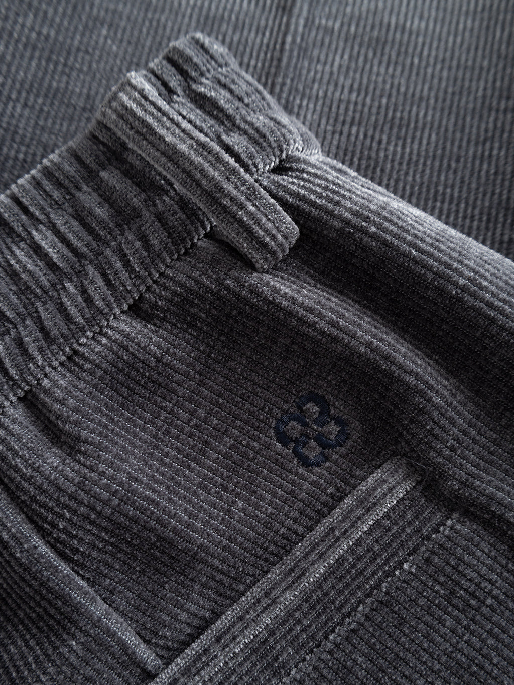 Close-up of Gabba’s GABmonza Cropped Tasca Fune Pant in Turbulence Blue, featuring dark gray ribbed jersey corduroy, an elastic waistband, and a small embroidered flower-like detail near the pocket.