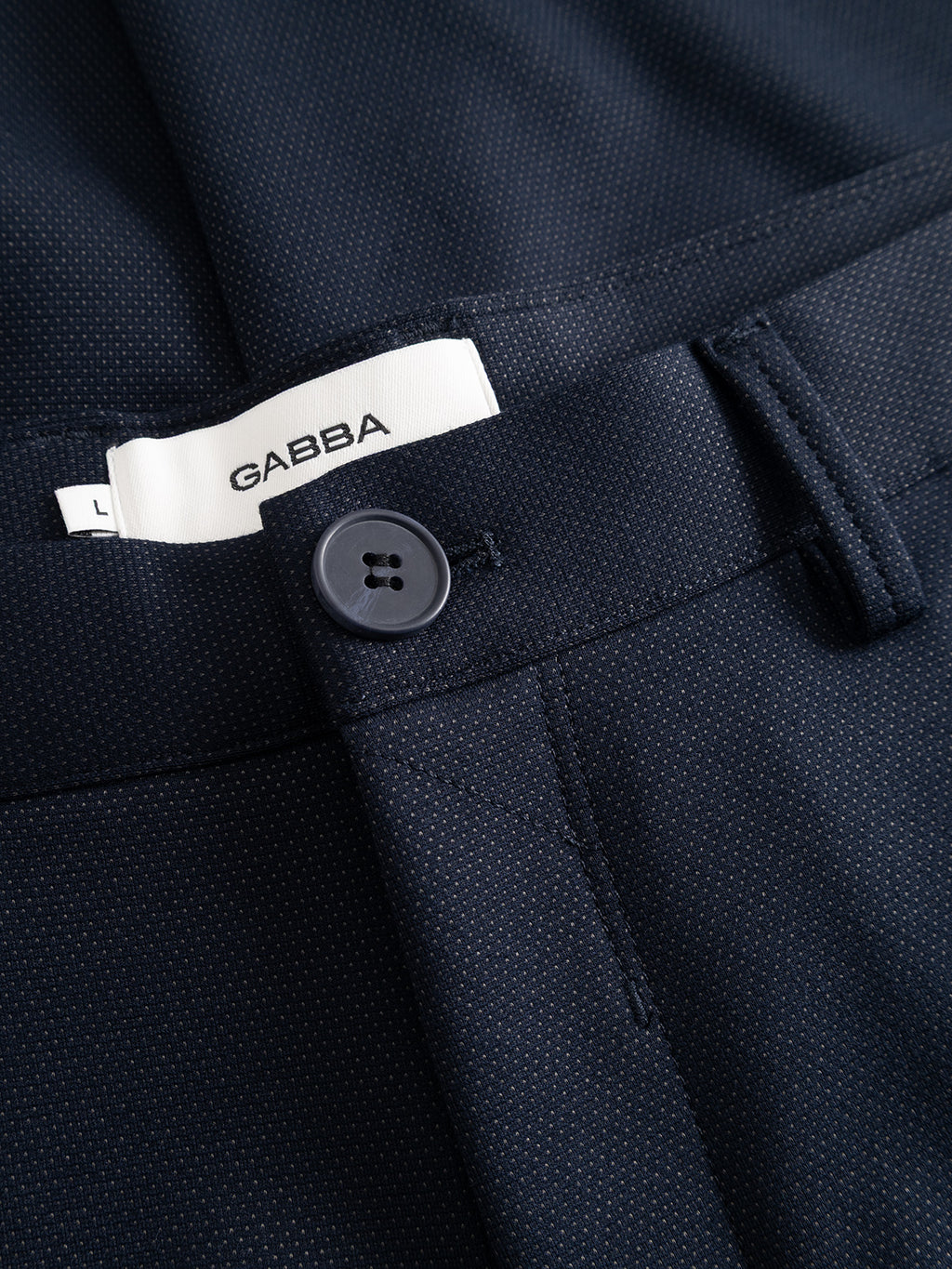 Close-up of the GABpaul Dotty Pant in navy by Gabba, showcasing a button closure, belt loops, and a visible white brand label. The slim chino fit is crafted from fabric with a refined dotted micro-pattern weave for added detail.