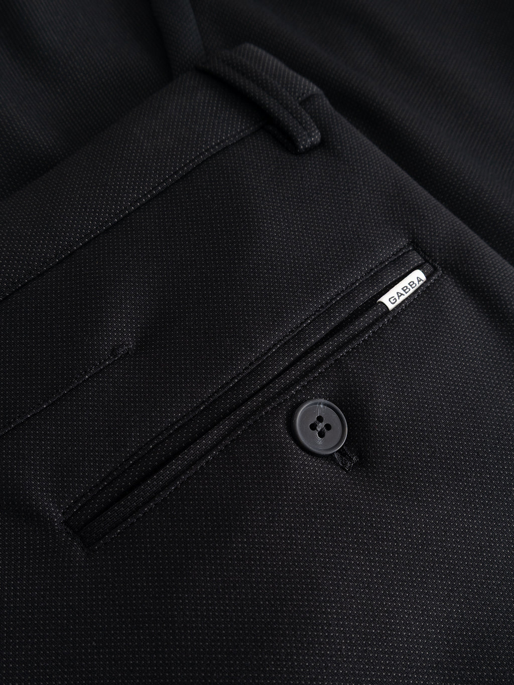 Close-up of the GABpaul Dotty Pant - Meteorite by Gabba, showcasing a black dress pant pocket detailed with a small white Gabba label, a zipper, and a black button. The superstretch slim chino features a refined micro-pattern weave for subtle texture.