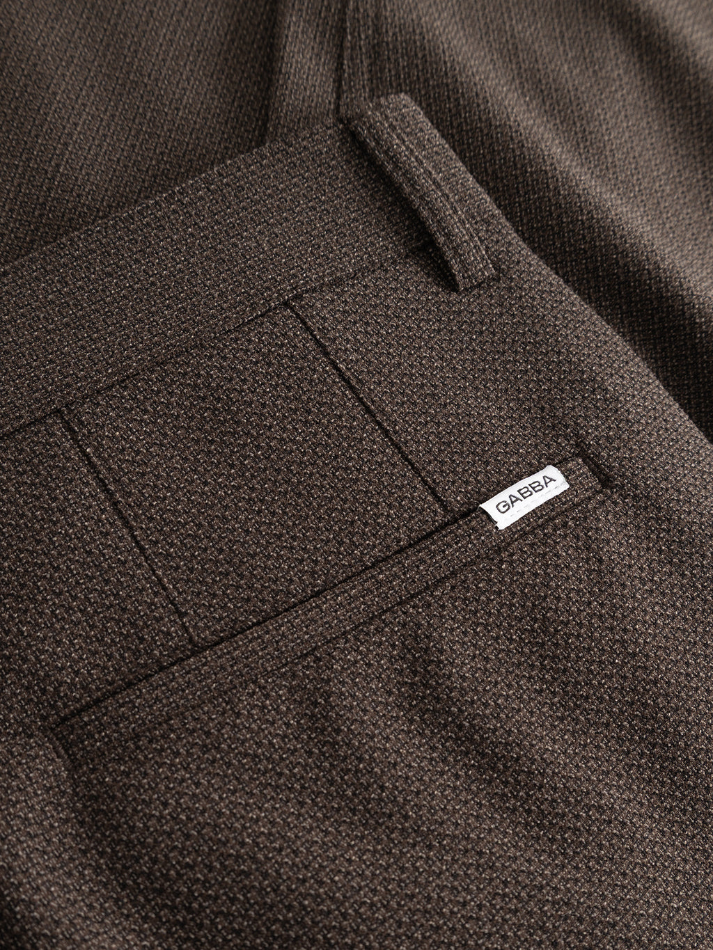 Close-up of the GABjoe-pas Rubin Pant in Brown Slate by Gabba, showcasing its tapered fit and visible back pocket with a white GARSEA label. These tailored trousers are crafted from textured dobby fabric with a fine, subtle pattern.