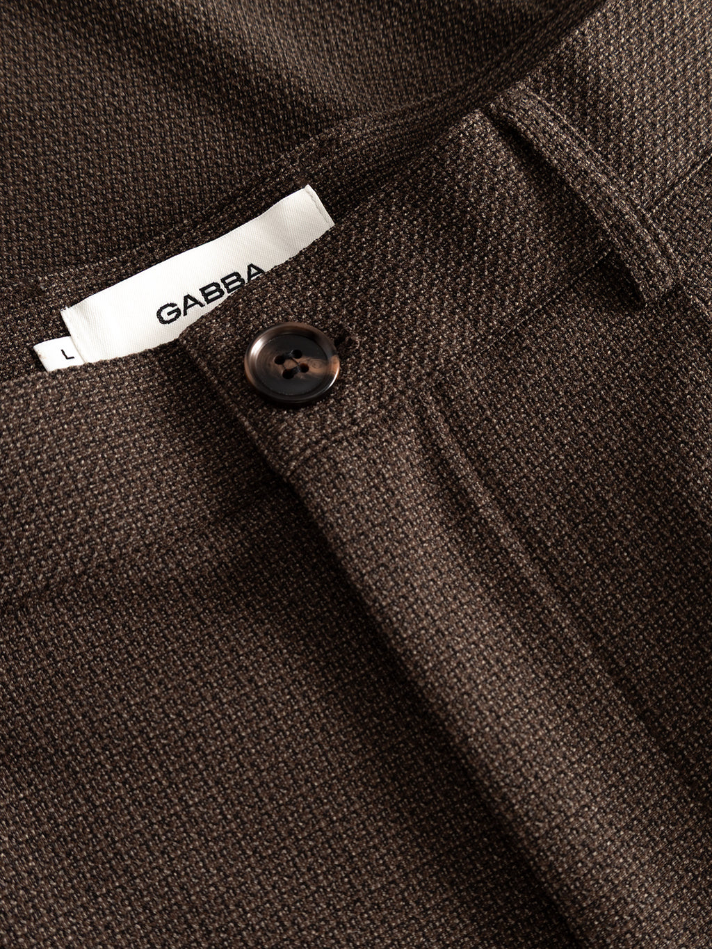 The GABdavid Rubin Pant in Brown Slate by Gabba showcases brown textured trousers made from stretch dobby fabric, highlighted with a close-up of the waistband featuring a black button, belt loop, and a distinctive white GABBA L label. These contemporary chino-style pants offer a loose tapered fit for modern comfort and versatility.