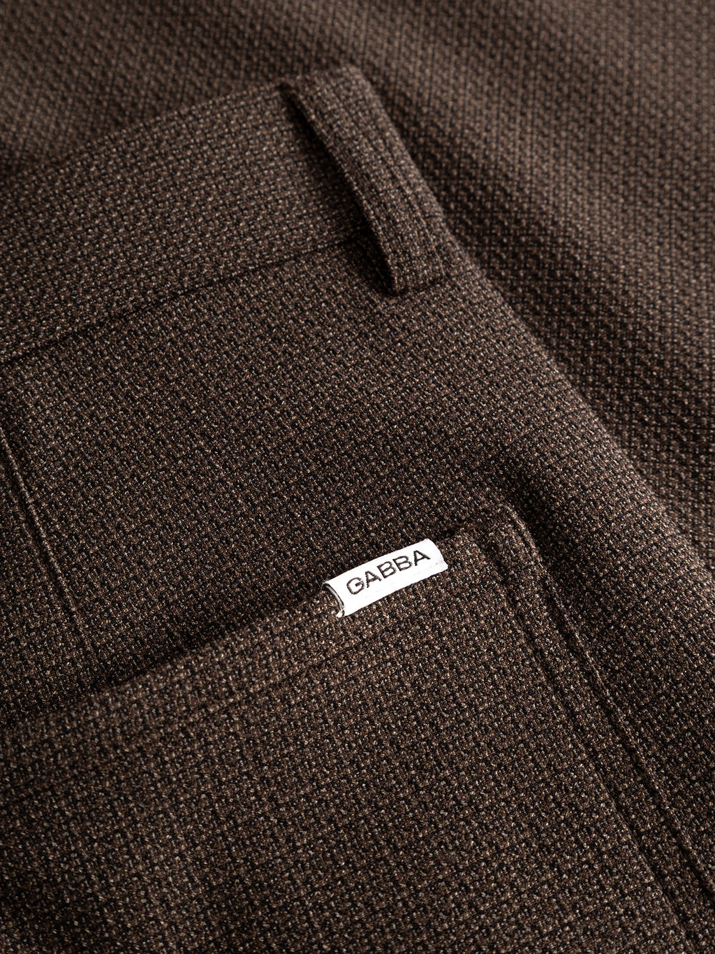Close-up of a textured brown stretch dobby fabric with a pocket. A small white label on the pocket reads Gabba. The visible weave pattern showcases the contemporary chino look and highlights the detailed stitching of the GABdavid Rubin Pant in Brown Slate by Gabba.