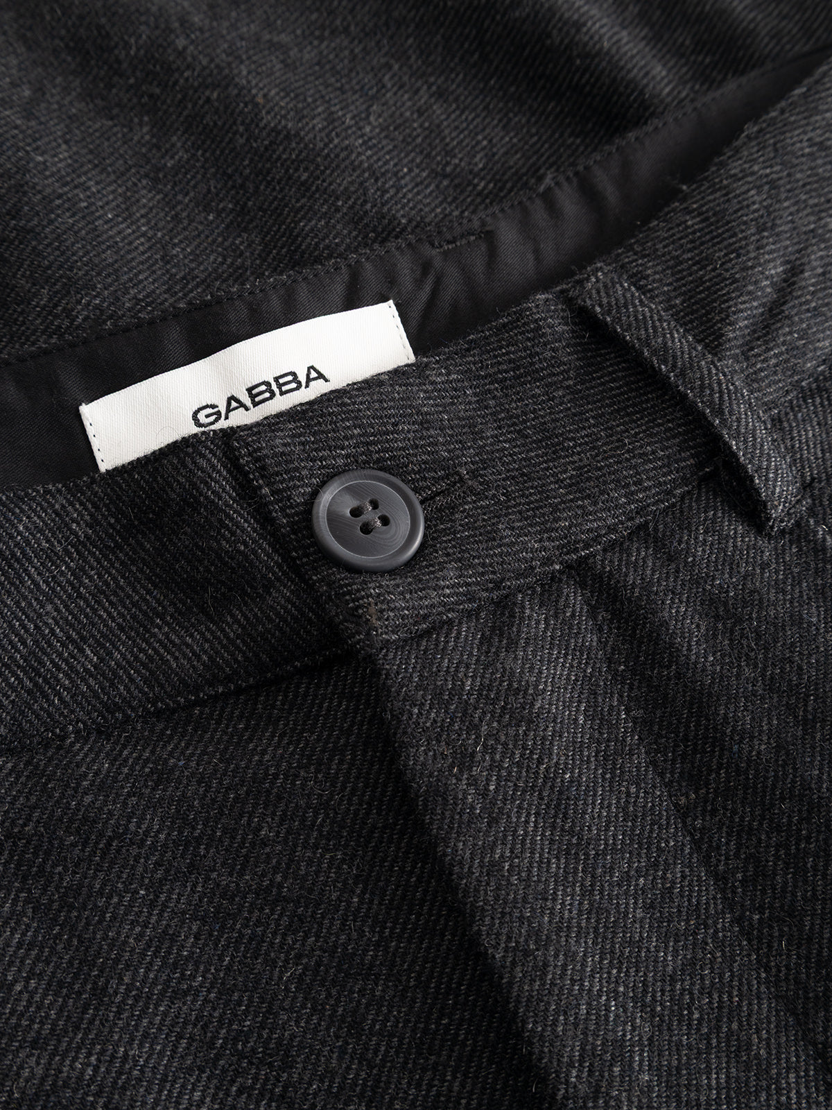Close-up of the dark grey GABdavid Twilight Pant by Gabba, featuring a black button and an interior white label with the Gabba logo sewn into the waistband. Crafted from a wool-blend twill fabric with a subtle textured pattern, these contemporary chinos offer a loose tapered fit for modern comfort.