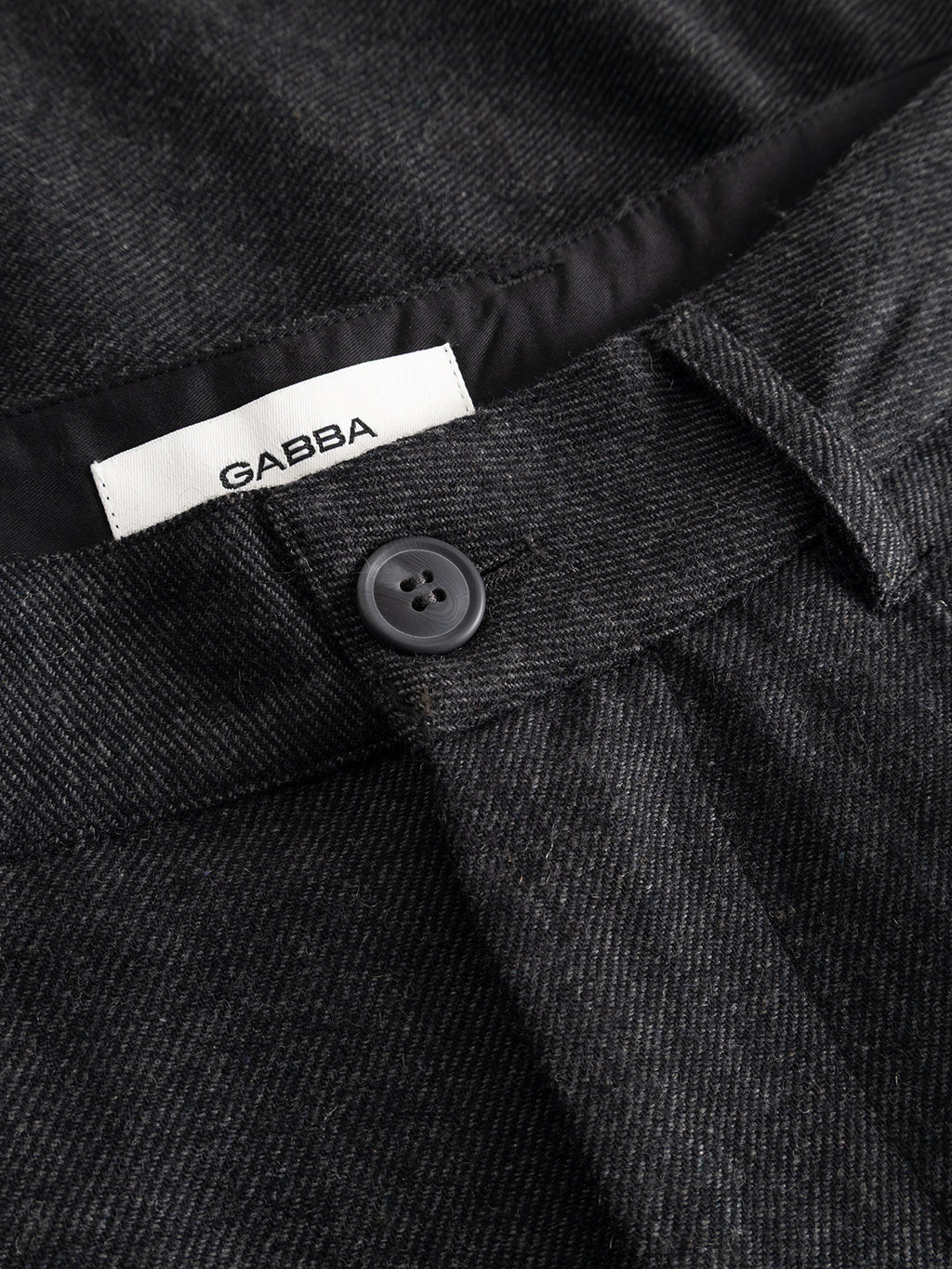 Close-up of the dark grey GABdavid Twilight Pant by Gabba, featuring a black button and an interior white label with the Gabba logo sewn into the waistband. Crafted from a wool-blend twill fabric with a subtle textured pattern, these contemporary chinos offer a loose tapered fit for modern comfort.