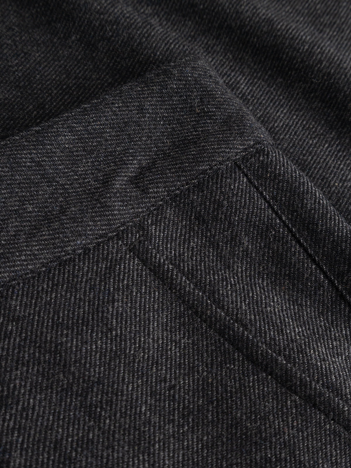 Close-up of the dark grey wool-blend twill fabric used in the Gabba GABdavid Twilight Pant, featuring a visible diagonal weave pattern and the corner of a sewn pocket. The texture and stitching highlight the contemporary design of these trousers, characterized by their loose tapered fit.
