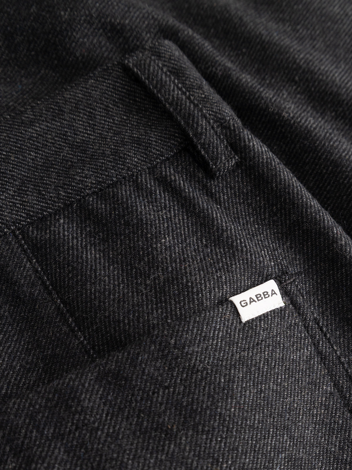 Close-up of a black textured wool-blend twill fabric pocket on the Gabba GABdavid Twilight Pant in Dark Grey, featuring a small white tag with the Gabba logo sewn near the seam, highlighting the refined details characteristic of contemporary chino styles.