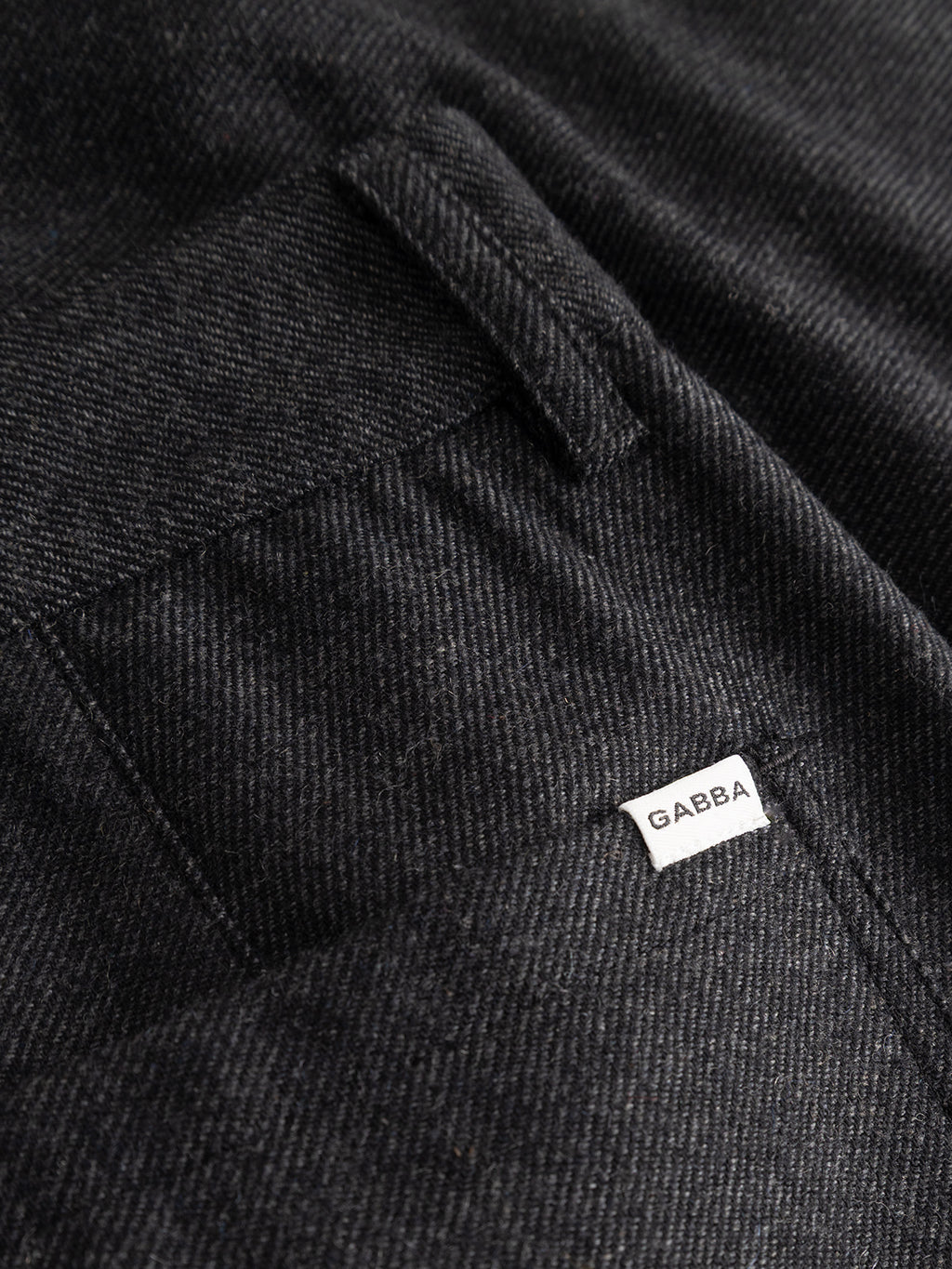 Close-up of a black textured wool-blend twill fabric pocket on the Gabba GABdavid Twilight Pant in Dark Grey, featuring a small white tag with the Gabba logo sewn near the seam, highlighting the refined details characteristic of contemporary chino styles.