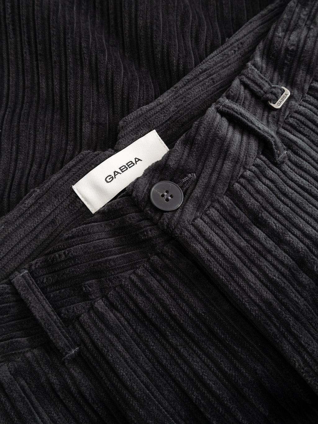Close-up of the GABfirenze Cropped Riglia GOTS Pant in Dark Grey by Gabba, featuring pleated detailing, a button closure, waistband, belt loops, and a white Gabba label. Crafted from organic cotton corduroy with a ribbed texture, these pants offer a relaxed fit and cropped length for modern style.