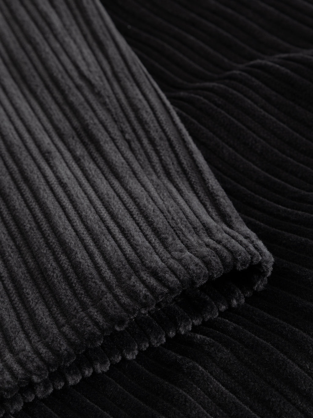 Close-up detail of the Gabba GABfirenze Cropped Riglia GOTS Pant in Dark Grey, featuring black ribbed velvet fabric reminiscent of organic cotton corduroy, with visible parallel vertical ridges and a soft, plush texture where one section overlaps another.