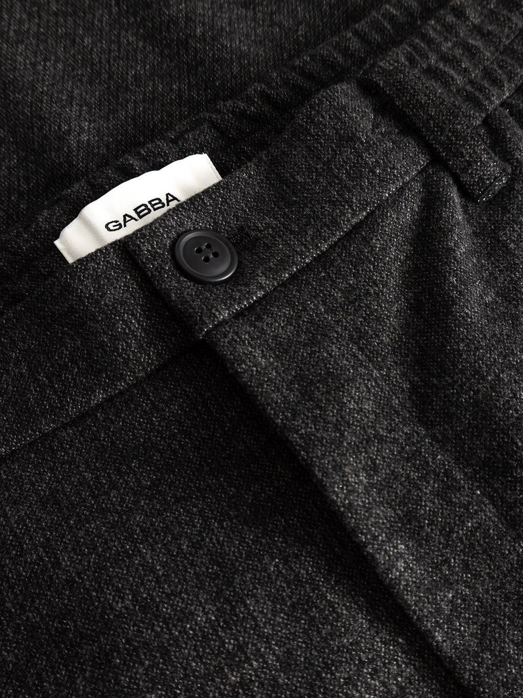 Close-up of the GABmonza Cropped Tasca Zaffe GRS Pant in dark grey by Gabba, showcasing its wool-look textured finish, a black button, and a visible white Gabba label near the waistband.