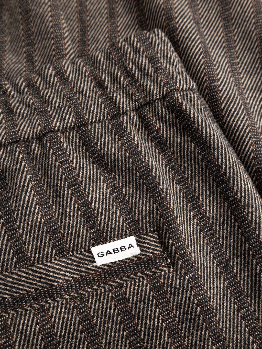 Close-up of the brown slate herringbone-patterned fabric of the Gabba GABnapoli 19 Zapies Pant, featuring a white tag with “Gabba” sewn on and highlighting the wool-look stripe detail characteristic of these regular straight fit trousers.