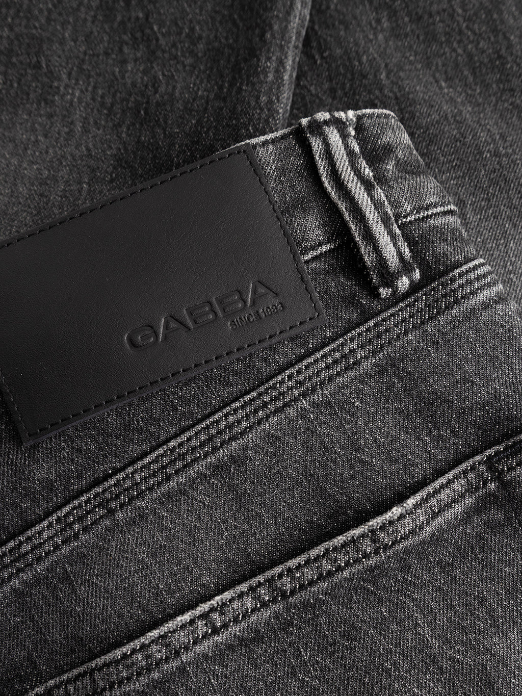 Close-up of the GABcarl Blackrow DH0011 Jeans in black denim from Gabba, designed in a loose tapered fit with a washed black finish. These jeans showcase visible white stitching and a black leather waistband patch embossed with the Gabba brand name and “since 1983.” Crafted from comfort stretch black denim, they offer ease of movement and all-day comfort.