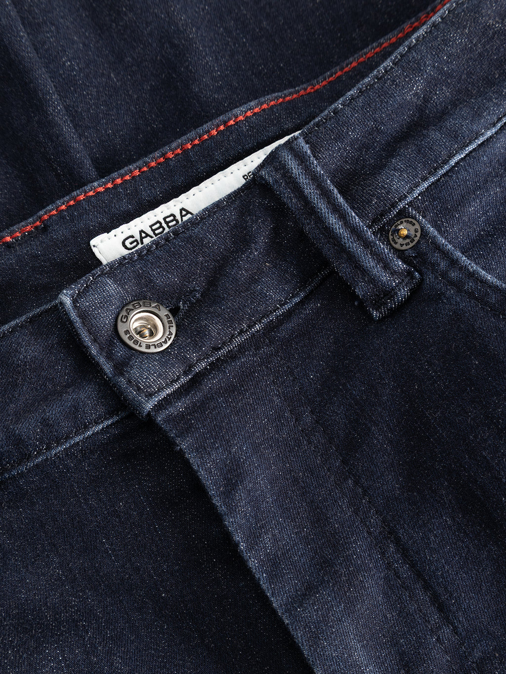 Close-up of the GABjones Vanew Rinse Jeans by Gabba in dark blue denim, featuring a slim straight fit with a visible button, belt loop, zipper area, distinctive red stitching, and a partially visible white label displaying the Gabba logo. Made from premium ISKO superstretch denim for superior comfort and flexibility.