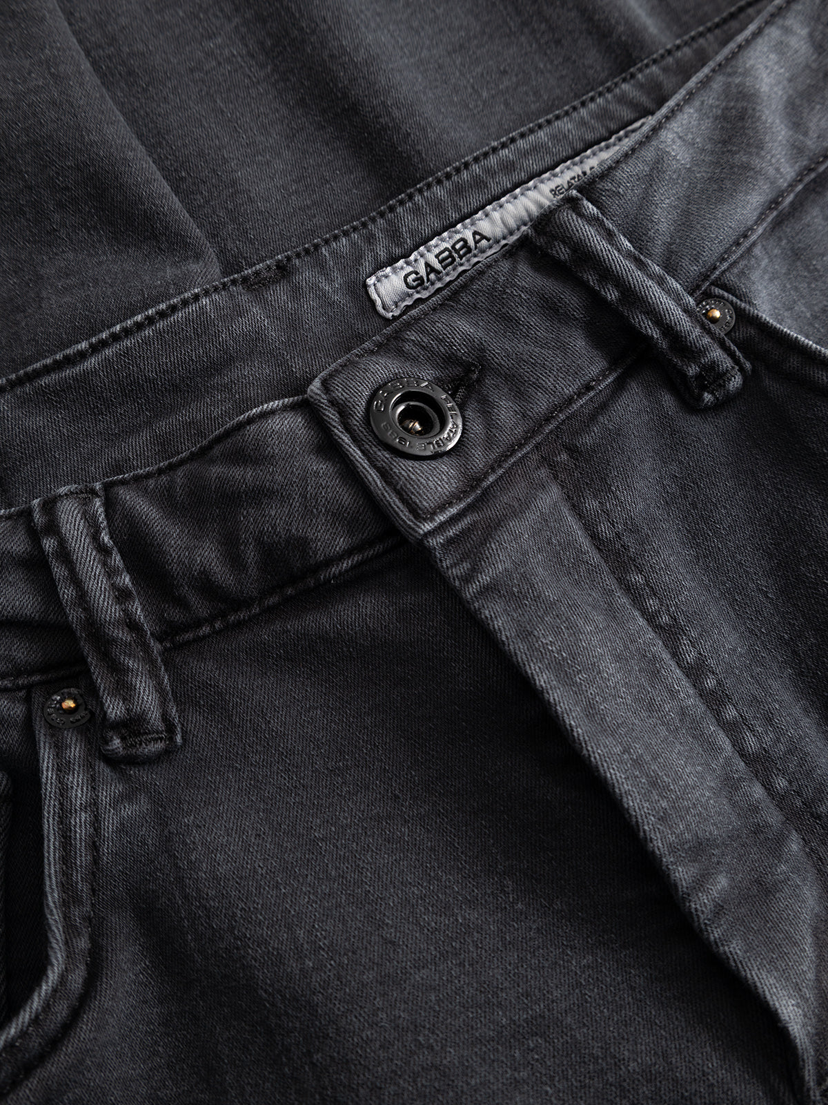 Close-up of the GABjones Hesky Jeans by Gabba in black denim, featuring high-stretch, dark gray fabric with a visible waistband, button, belt loops, zipper, and a label that reads Gabba. The slightly worn and textured denim gives these modern slim straight fit jeans a contemporary look.