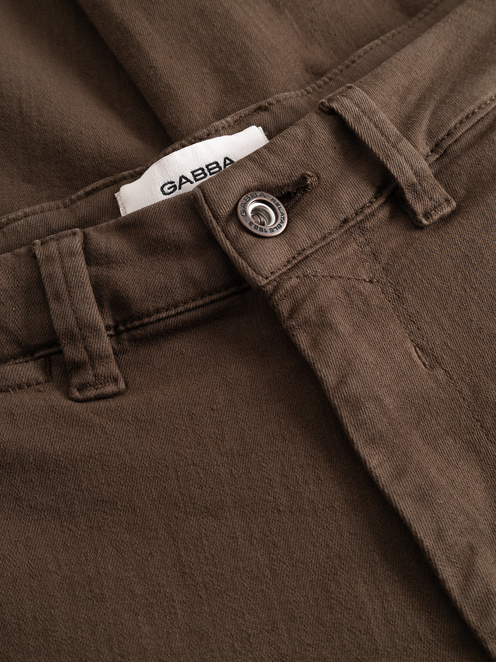 Close-up of the GABpaul Hesky Pant in brown by Gabba, featuring a metal button, belt loops, and a visible white label with the Gabba brand name. Made from garment-dyed denim or twill in a slim chino fit, these pants combine modern style with comfortable wear.