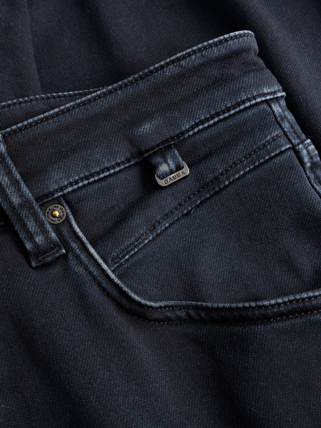 Close-up of a black denim jeans pocket with visible stitching, a metal rivet, and a small metallic tag labeled Gabba above the pocket. Crafted from premium indigo denim in a dark blue wash, the GABalex Cropped Torino RS2106 Jeans by Gabba feature a relaxed tapered fit for added comfort and style.