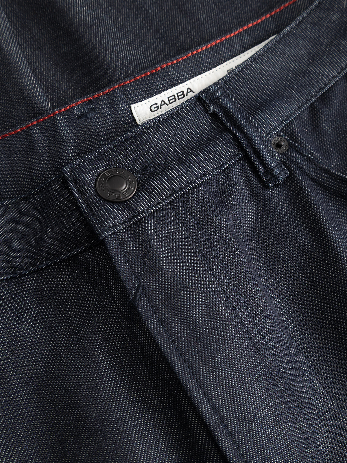 Close-up of GABcarl Anark Stone Raw Jeans by Gabba in dark blue denim, showcasing a black button, belt loop, and a white label with Gabba branding. Red stitching accents the waistband, highlighting the premium ISKO denim fabric in a loose tapered fit.