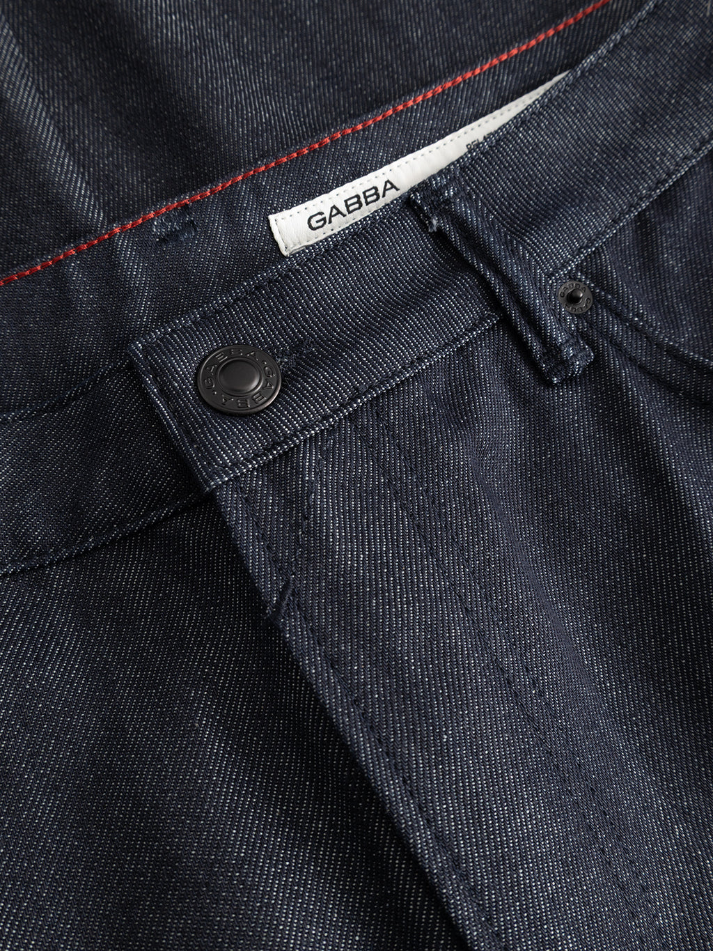 Close-up of GABcarl Anark Stone Raw Jeans by Gabba in dark blue denim, showcasing a black button, belt loop, and a white label with Gabba branding. Red stitching accents the waistband, highlighting the premium ISKO denim fabric in a loose tapered fit.