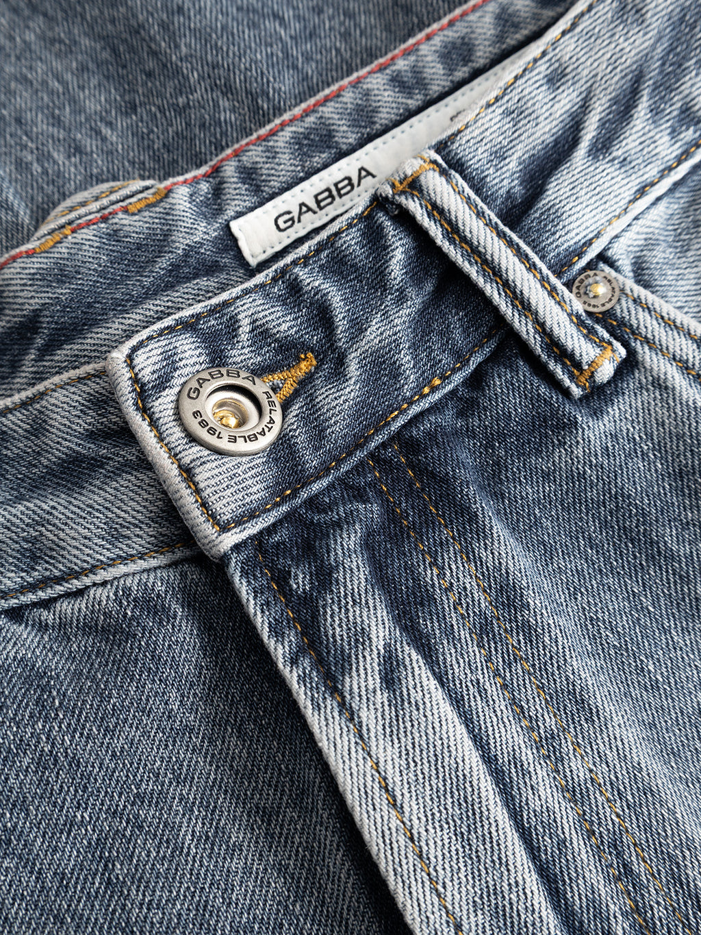 Close-up of the GABcarl Stone RS2103 Jeans by Gabba, featuring mid-blue denim with a metal button, yellow stitching, and a white label with the Gabba logo. Crafted from premium ISKO denim in a mid-blue stone wash, the fabric displays visible fading and texture.