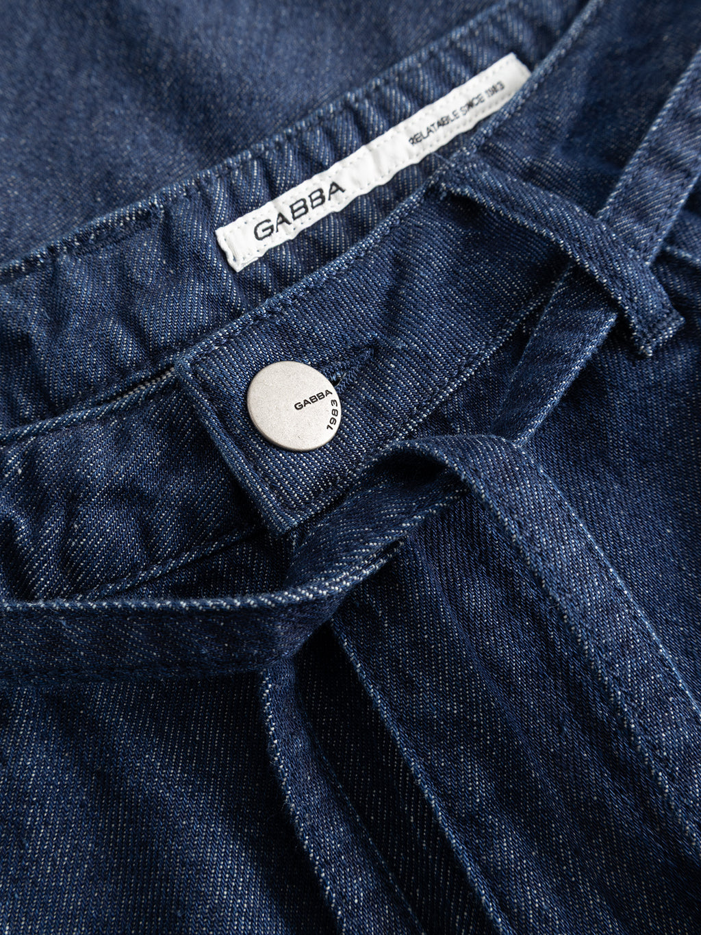 Close-up of the GABnoah Tona Rinse Jeans in dark blue denim, crafted from a rigid indigo organic cotton blend. The jeans feature visible stitching, a silver button, and a white Gabba label sewn onto the waistband, showcasing their loose straight fit design.