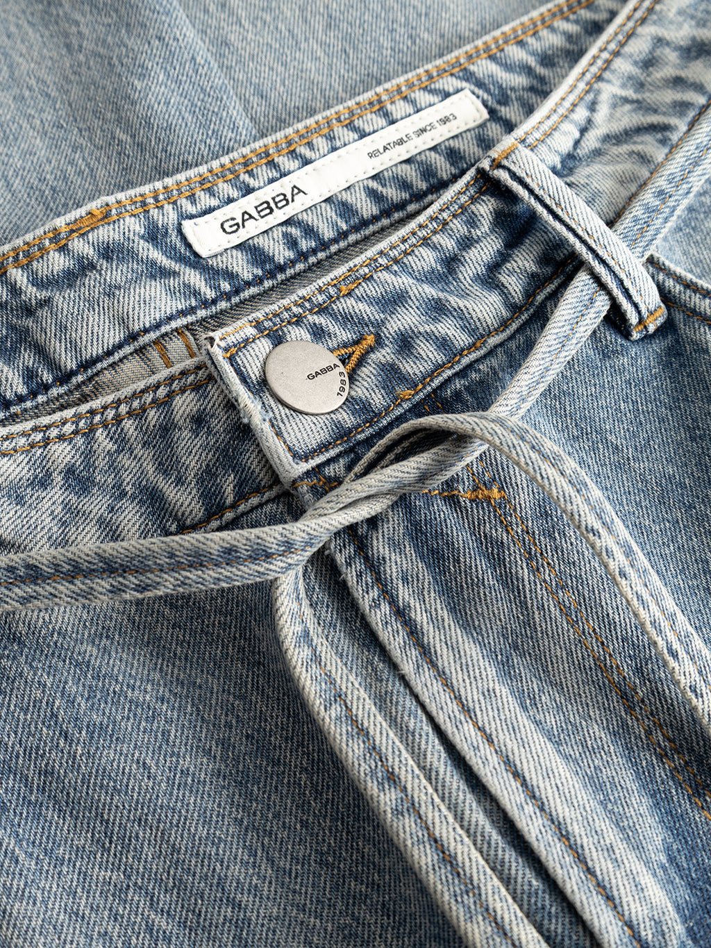 Close-up of the GABmax Tona RS2100 Jeans in mid blue denim by Gabba, featuring a metal button, belt loops, and a white inner waistband label reading Gabba. Orange stitching along the seams accentuates the craftsmanship of these modern straight fit jeans.