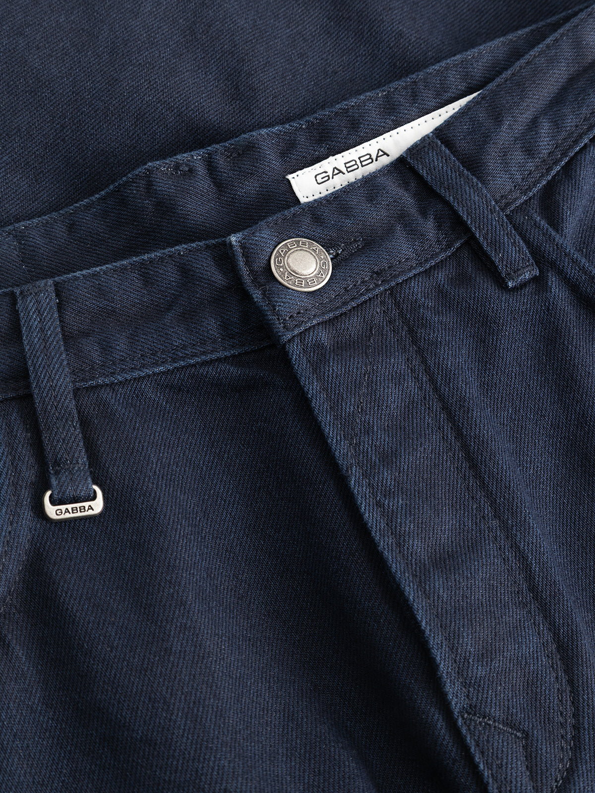 Close-up of the GABsenna Denia Rinse Jeans in dark blue denim, featuring a metal button, belt loops, and both a small metal tag and fabric label displaying the Gabba brand name. These relaxed fit jeans are crafted from premium blue-black denim for a blend of style and comfort.