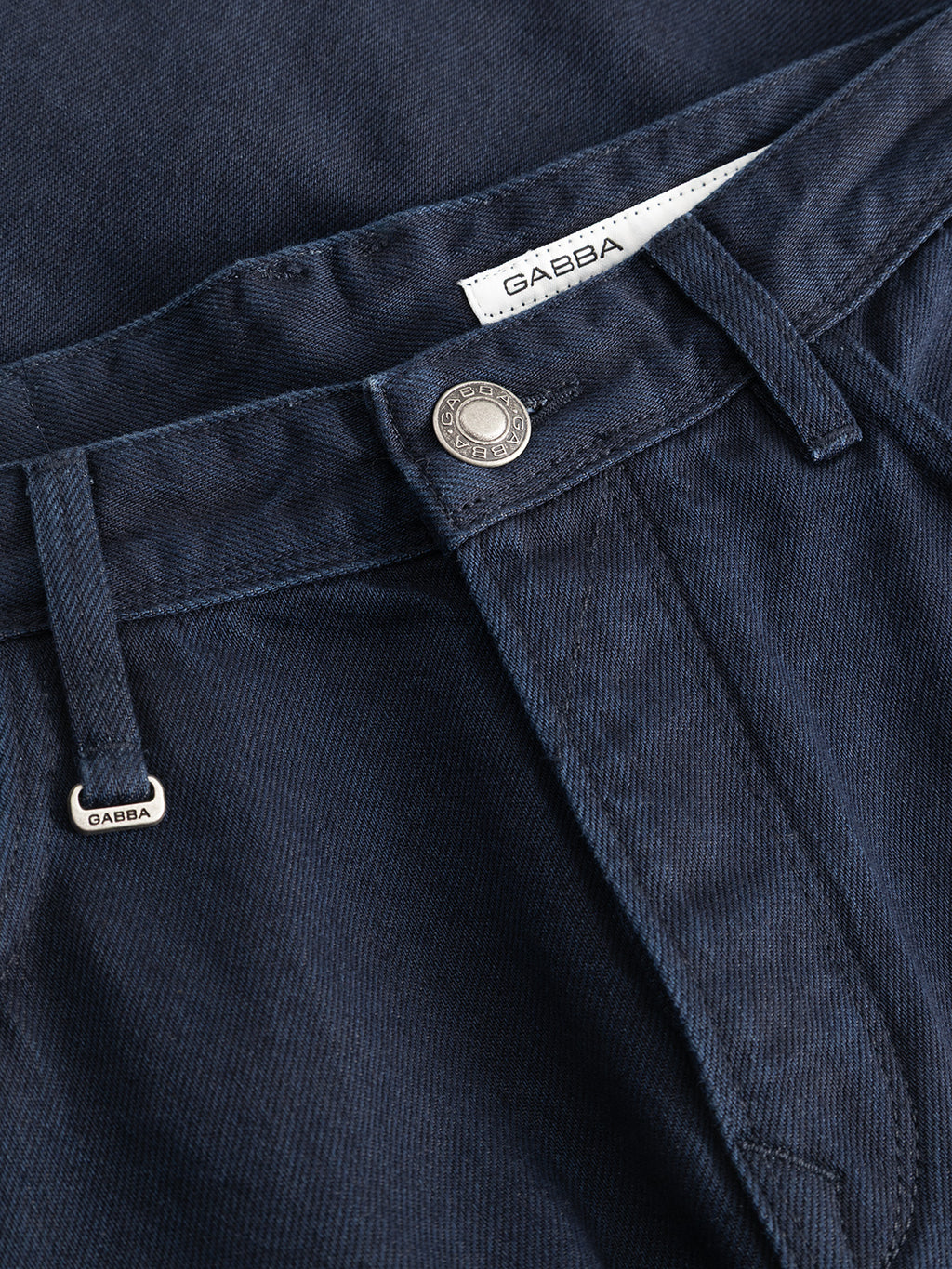Close-up of the GABsenna Denia Rinse Jeans in dark blue denim, featuring a metal button, belt loops, and both a small metal tag and fabric label displaying the Gabba brand name. These relaxed fit jeans are crafted from premium blue-black denim for a blend of style and comfort.