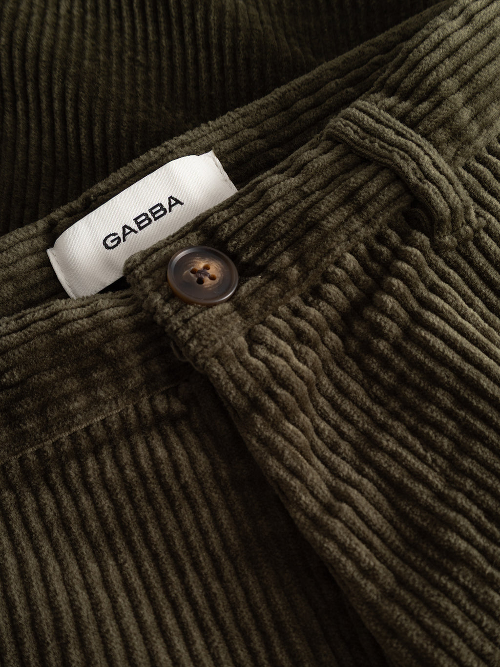 Close-up of Gabba GABjoe Nopleats Ibre Pant in Forest Night, featuring olive green stretch corduroy, a brown button, and a white GABBA label on the waistband, highlighting classic chino construction.