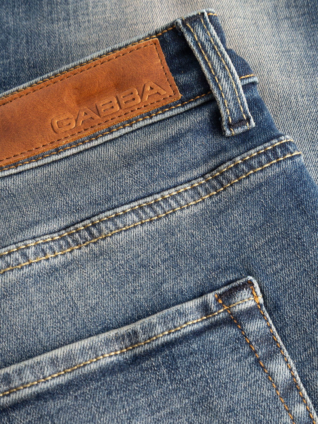 Close-up of the Gabba GABmarc Bekham DS0003 Jeans in mid-blue denim, featuring a brown leather patch embossed with the Gabba logo above the back pocket. The visible stitching and fabric texture emphasize the comfort stretch denim and regular fit design.