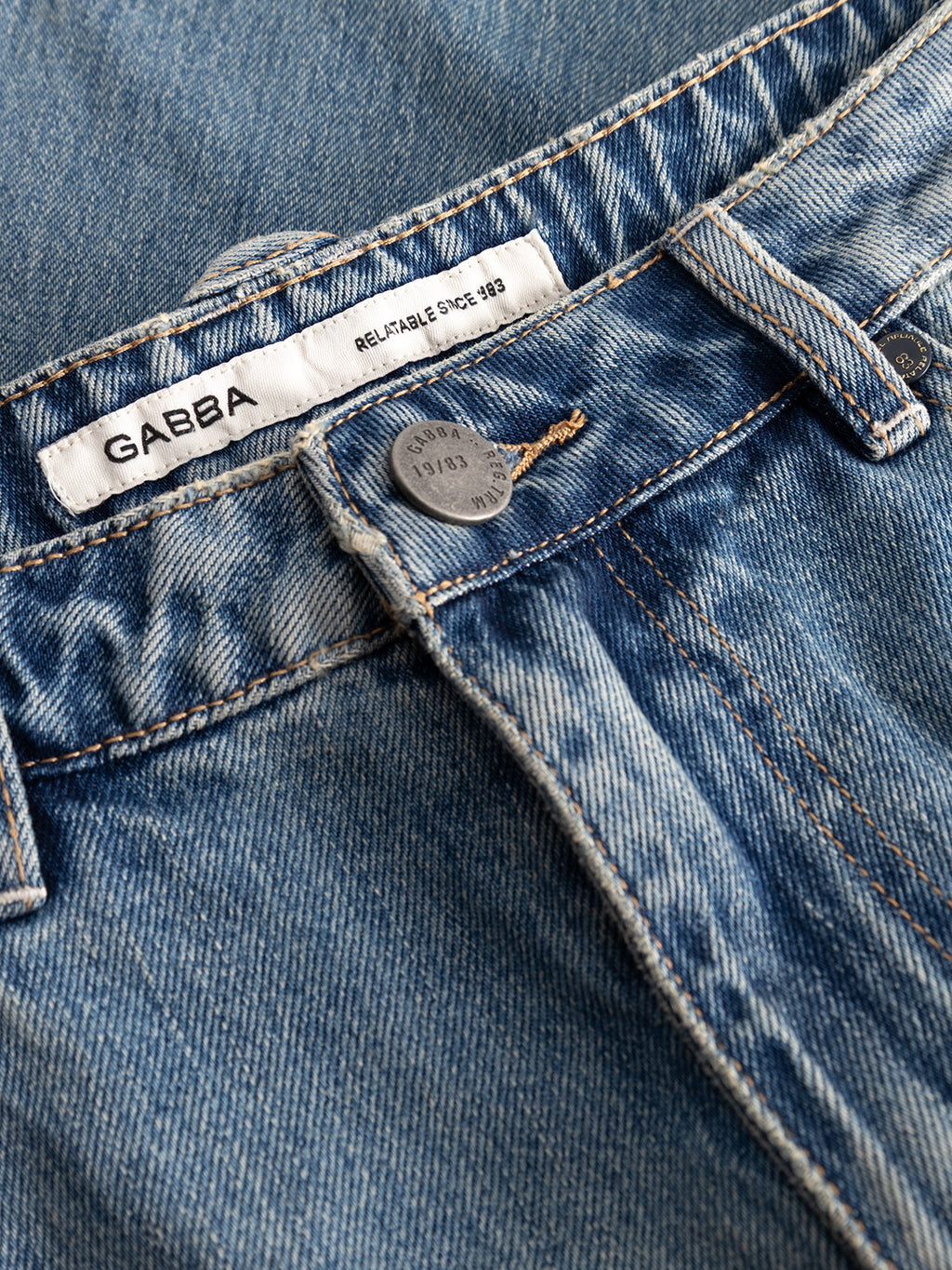 Close-up of GABcarl Bieffe GW0001 Jeans in mid-blue denim, showcasing the waistband, button, and a white label featuring the Gabba brand name along with “RELIABLE SINCE 1983.” Made from slub denim, these jeans display visible stitching, faded details, and offer a loose tapered fit for enhanced comfort and style.