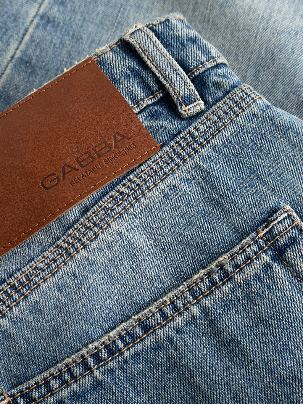 Close-up of the GABcarl Bieffe GW0001 Jeans in mid blue denim, featuring a brown leather patch on the waistband embossed with the Gabba brand name and the text “RELATABLE SINCE 1983.” Crafted from slub denim, these jeans showcase visible belt loop and stitching details, offering a relaxed yet refined loose tapered fit.