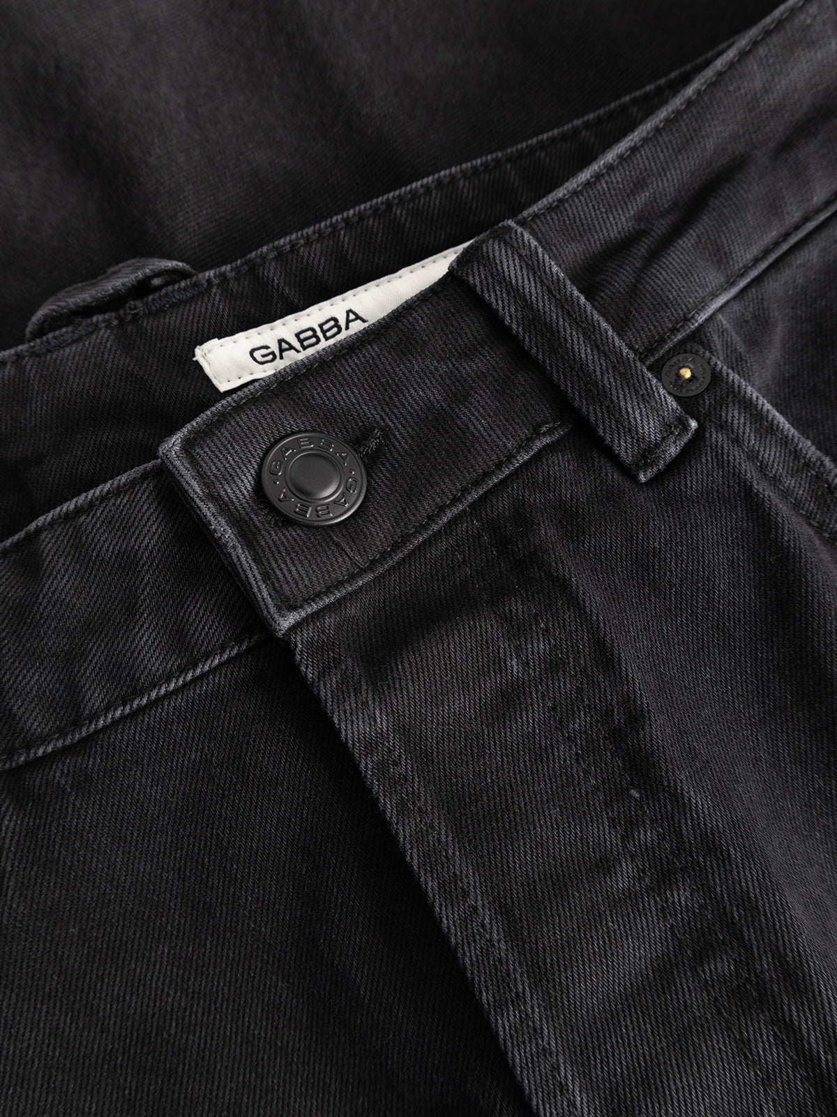 A close-up of the GABcarl Blaccom WW0035 Jeans in black denim highlights the waistband, a black button, detailed stitching, and a white label with the Gabba brand name partially visible. Crafted from relaxed denim for all-day comfort, these jeans offer a loose tapered fit that seamlessly combines style and ease.