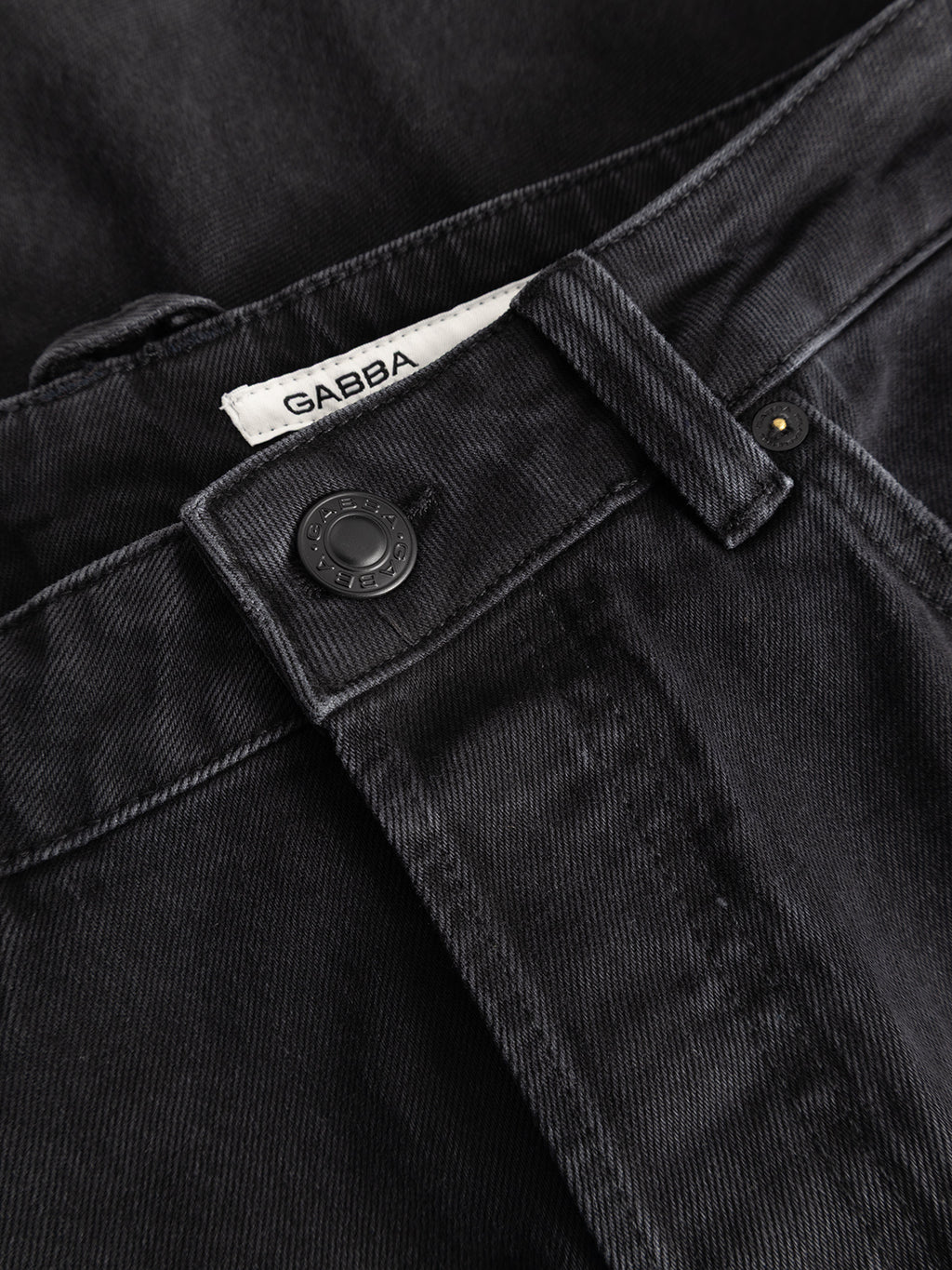 A close-up of the GABcarl Blaccom WW0035 Jeans in black denim highlights the waistband, a black button, detailed stitching, and a white label with the Gabba brand name partially visible. Crafted from relaxed denim for all-day comfort, these jeans offer a loose tapered fit that seamlessly combines style and ease.