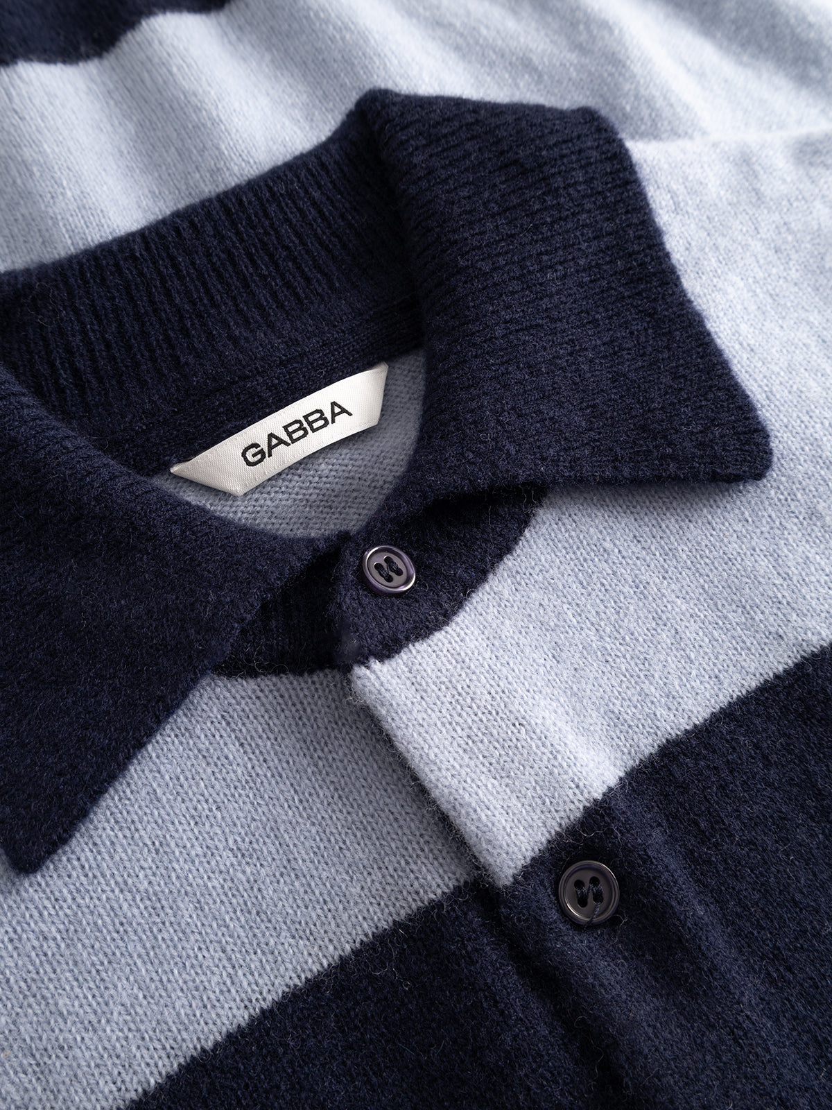 Close-up of the GABworren Polo Stripes Aico LS Knit - Navy Stripe, a men’s regular fit pullover polo sweater from Gabba, featuring navy blue and light gray stripes, a collared neckline with buttons, and an inner collar tag displaying the brand name.