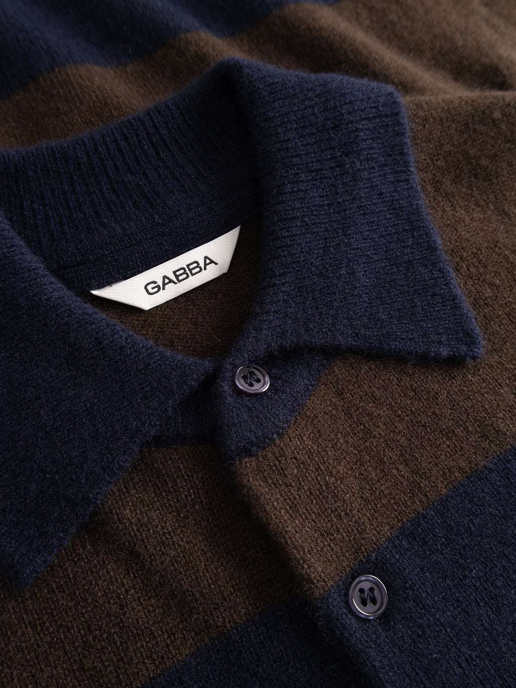 Close-up of the Gabba GABworren Polo Stripes Aico LS Knit in brown stripe, featuring navy and brown stripes, a collared neckline, and two dark buttons. The soft wool knit texture is clearly visible, and a white label inside the collar displays “GABBA.”.