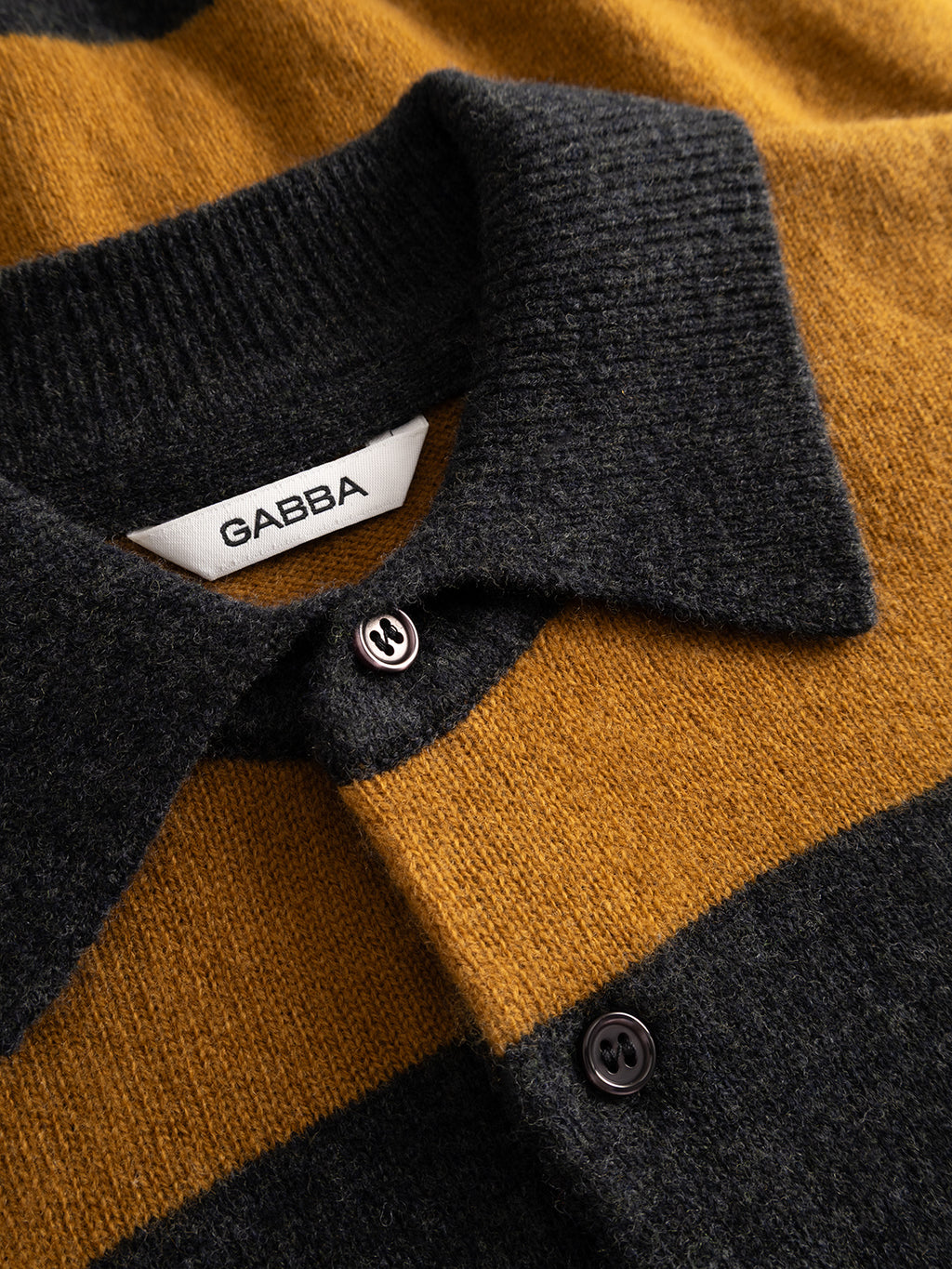 Close-up of the Gabba GABworren Polo Stripes Aico LS Knit sweater for men, featuring a regular fit, wide mustard yellow and dark gray stripes, black buttons, and a white label reading “Gabba.”.