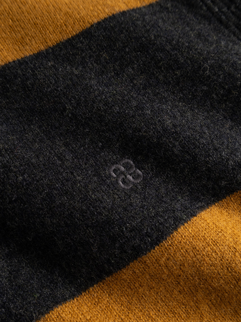 Close-up of the GABworren Polo Stripes Aico LS Knit - Yellow Stripes by Gabba, a men’s regular fit sweater crafted from soft wool with bold horizontal black and mustard yellow stripes. This knit features a small, subtle embroidered symbol in black thread on the black stripe.