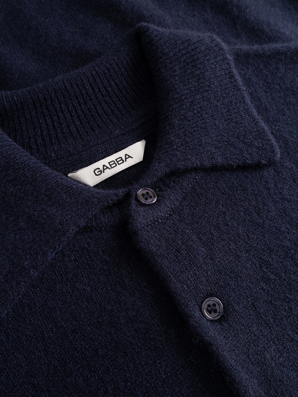 Close-up of the Gabba GABworren Polo Aico LS Knit in navy—a dark wool shirt featuring a collar, two dark buttons, and a visible white label inside the neck reading “Gabba.” This design draws inspiration from timeless knitwear, blending classic style with modern details for versatile wear.