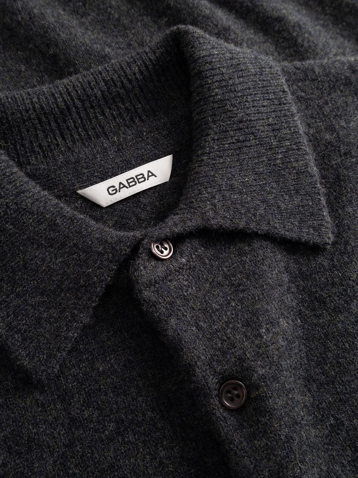 Close-up of the GABworren Polo Aico LS Knit in Dark Grey Mel.—a soft wool knit shirt featuring a ribbed polo collar and black buttons. The interior of the collar showcases a white Gabba label, emphasizing the refined details of this men’s regular fit sweater.