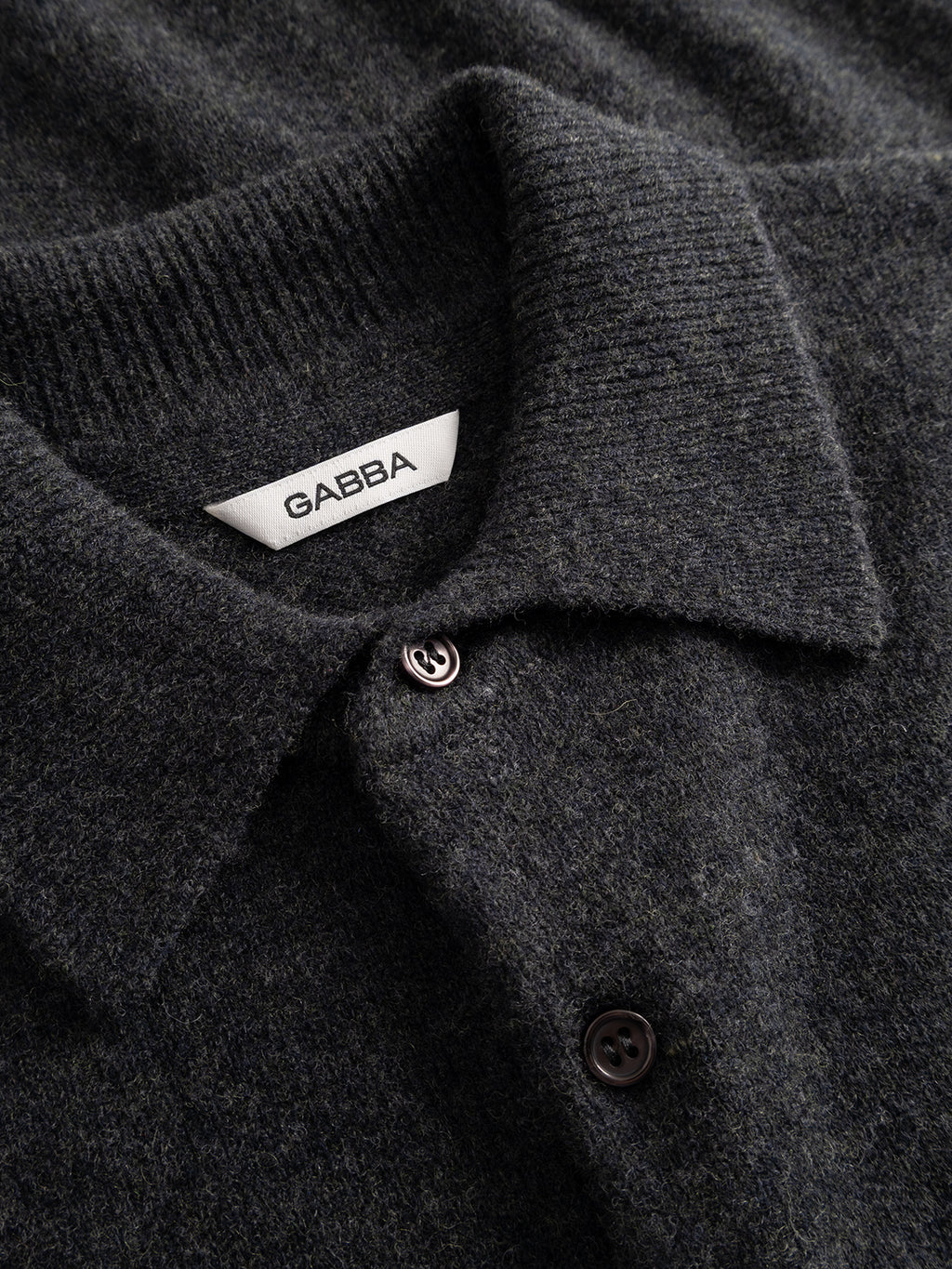 Close-up of the GABworren Polo Aico LS Knit in Dark Grey Mel.—a soft wool knit shirt featuring a ribbed polo collar and black buttons. The interior of the collar showcases a white Gabba label, emphasizing the refined details of this men’s regular fit sweater.